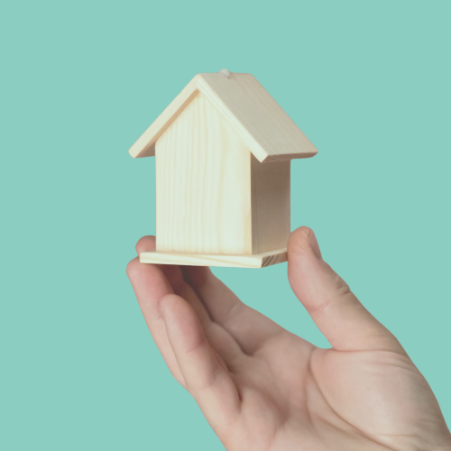 Casa pequeña de madera en mano sobre fondo azul Mano sosteniendo una casita de madera sobre un fondo azul